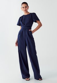 Mornarško modra kombinezon, kratki napihnjeni rokavi, ovit dizajn v pasu, širok silhueta hlač, izdelan iz gladkega materiala, ima sprednje žepe.