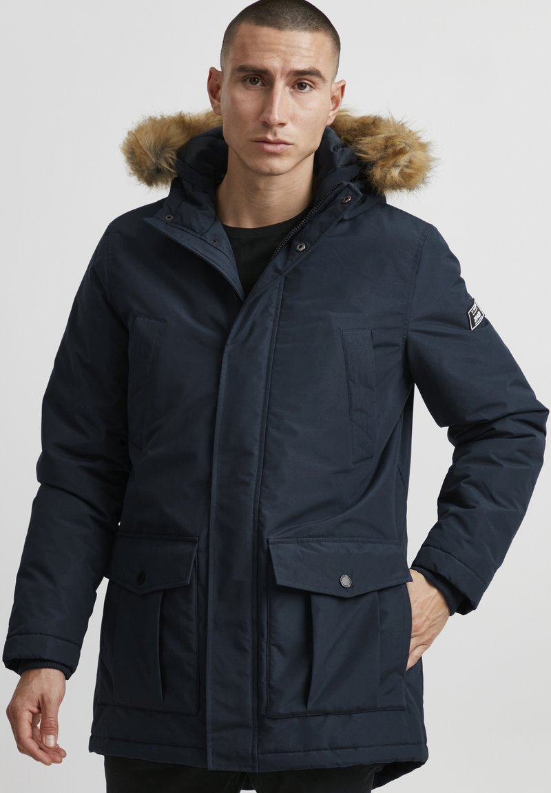 Marineblå parka med hette foret med faux pelasje, trykknapper, to frontlommer og et glatt, vannavstøtende stoff.