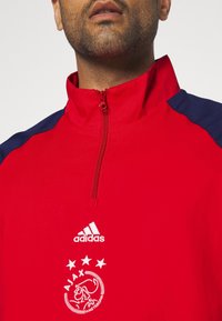 Chándal deportivo rojo con cuello de media cremallera, acentos en azul en los hombros, logo blanco de Adidas, emblema del Ajax de Ámsterdam y motivo de tres estrellas.