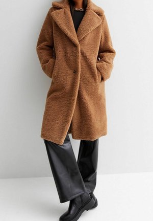 Mujer con un abrigo teddy marrón hasta la rodilla con solapas anchas, pantalones de cuero negros y botines negros, de pie con las manos en los bolsillos del abrigo.