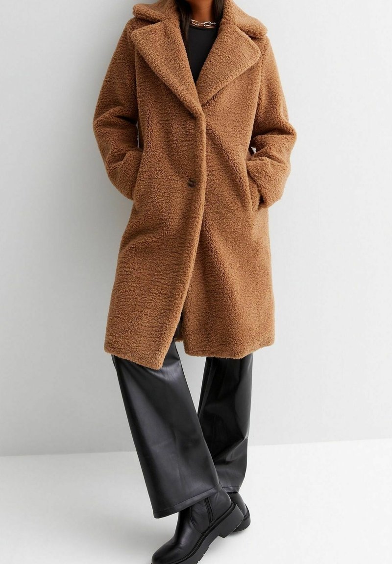 Femme portant un manteau teddy marron à larges revers, longueur genoux, un pantalon en cuir noir et des bottines noires, debout les mains dans les poches du manteau.