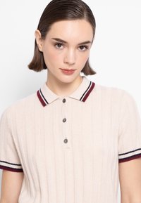 Polo di colore beige chiaro con texture a coste, colletto a righe rosso scuro e blu navy, e chiusura a tre bottoni. Maniche corte con accenti abbinati.