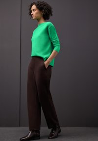 Pull en tricot vert à col rond, associé à un pantalon marron foncé. Le mannequin porte des bottines brillantes et a les cheveux courts et bouclés.