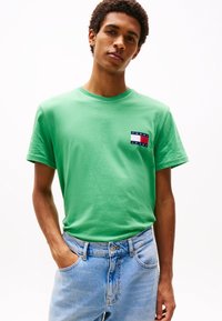 Young man wearing a green Tommy Jeans t-shirt and light blue jeans, standing with one hand in pocket against a white background.