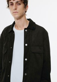 Zwarte suède button-up jas met zilveren drukknoop hardware, twee borstzakken en een notch kraag, gedragen over een wit T-shirt.