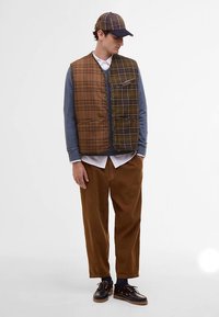 Hnědá károvaná prošívaná vesta s předními kapsami, nošená přes modrou košili s dlouhým rukávem a bílým límečkem, v kombinaci s hnědými kalhotami a tmavými botami.