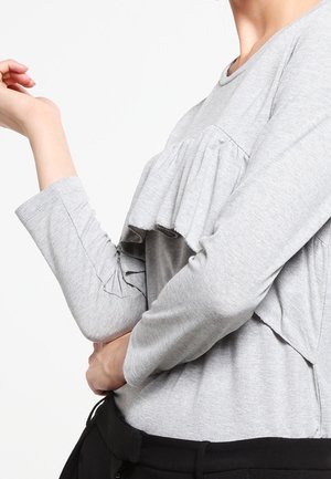 Personne portant un haut gris clair à manches longues avec un détail à volants sur la poitrine et un pantalon noir, bras plié et main près du visage.