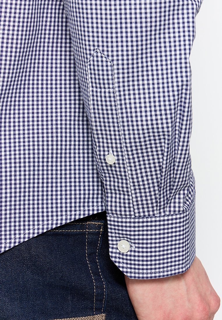 Camisa de cuadros en azul y blanco con puños abotonados, que presenta un diseño de puño puntiagudo y una textura de algodón suave. Combinada con jeans de mezclilla oscura.