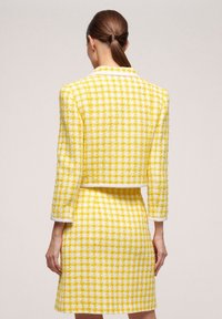 Žlutý a bílý károvaný tweedový kabátek s krátkým střihem, jednobarevným bílým lemem a třičtvrtečními rukávy, hranatý s odpovídající sukní.