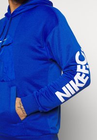 Hoodie bleu avec une poche kangourou, un tissu texturé et un grand logo blanc "NIKE" imprimé sur la manche.