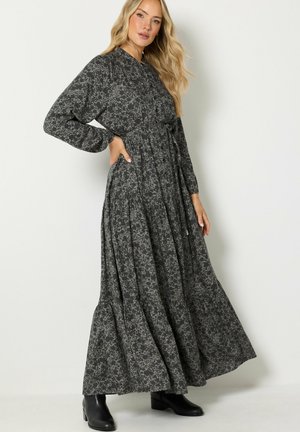 Woman with long blonde hair wearing a long-sleeve dark floral maxi dress and black ankle boots standing against a plain background.