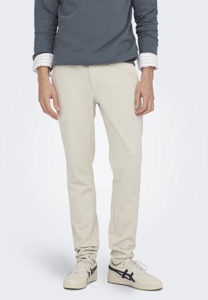 Mann trägt beige Slim-Fit-Hosen, marineblaue Sweatshirt mit aufgerollten Ärmeln und weiße Sneakers mit schwarzen Streifen, steht auf weißem Hintergrund.