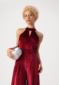 Burgundinė aksominė halter jumpsuit su raktų skylės iškirpte ir klostuotomis kelnėmis, derinama su sidabriniu, apvaliu rankine.