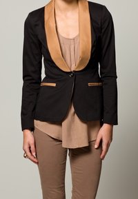Blazer noir avec un col en satin beige et des accents, doté de deux poches avant et d'une fermeture à un bouton, associé à un haut marron clair.