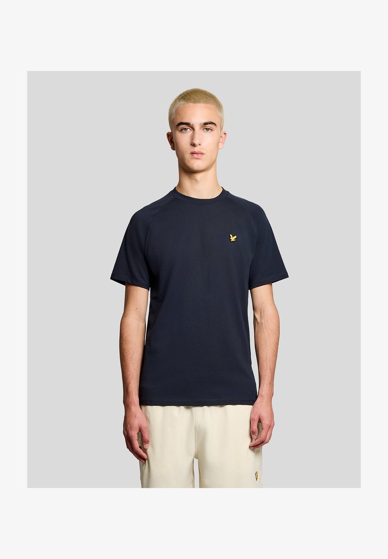 Jeune homme aux cheveux blonds courts portant une chemise à manches courtes bleu marine et un pantalon beige, debout devant un fond gris uni.