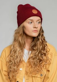 Bordowa dziana czapka z prążkowanym wzorem i okrągłą skórzaną naszywką w kolorze beżowym. Model nosi żółtą kurtkę i białą koszulkę, a jego kręcone włosy są widoczne.