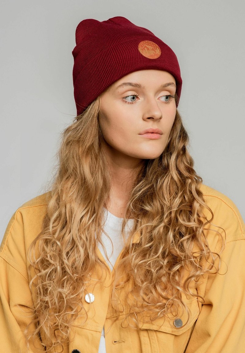Bordowa dziana czapka z prążkowanym wzorem i okrągłą skórzaną naszywką w kolorze beżowym. Model nosi żółtą kurtkę i białą koszulkę, a jego kręcone włosy są widoczne.