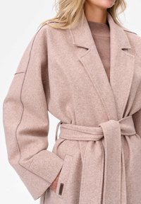 Beżowy płaszcz z wełny, w luźnym kroju, z szerokimi klapami i paskiem do wiązania. Posiada przeszycia oraz kieszenie z akcentem w postaci metki.