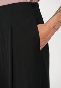 Pantalones negros a medida con una textura suave, que cuentan con un bolsillo lateral y un cierre de cremallera discreto, diseñados para un ajuste estructurado.