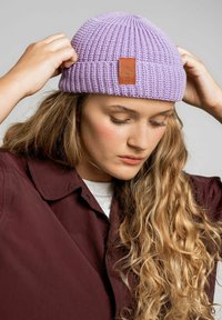 Fioletowa dziana czapka beanie z prążkowaną teksturą i skórzaną łatką z logo. Noszona przez osobę, która ją poprawia, w połączeniu z bordową koszulą z kołnierzykiem.