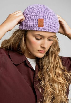 Fioletowa dziana czapka beanie z prążkowaną teksturą i skórzaną łatką z logo. Noszona przez osobę, która ją poprawia, w połączeniu z bordową koszulą z kołnierzykiem.