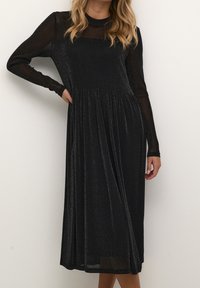 Svart glittrande klänning med långa genomskinliga ärmar, en figurnära bodice och en utsvängd kjol. Tyget har en subtil glans och mjuk textur.