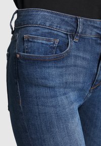 Acercamiento de unos pantalones vaqueros azules que muestran el bolsillo delantero, la ansa del cinturón y la costura detallada en una persona que lleva una camiseta negra.