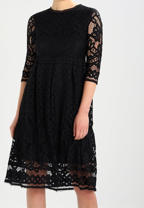 Robe noire en dentelle longueur genou avec manches trois-quarts, motifs floraux et géométriques, et col rond portée par une personne.