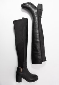 Bottes noires montant jusqu'au genou avec un design ajusté, fabriquées en similicuir lisse et tissu, dotées d'une fermeture éclair latérale et d'un talon épais.