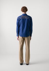 Takkipaita tummansinisestä denim-kankaasta, jossa on keltainen "Harlem" -brodeeraus selässä. Paidassa on pitkät hihat ja napit hihansuissa, yhdistettynä beigeihin housuihin.