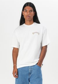 Hvid bomulds t-shirt med korte ærmer, der har et gult og sort grafisk logo på venstre bryst. Casual fit, parret med blå denim shorts.