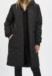 Gepadded schwarzer langer Mantel mit Kapuze, ausgestattet mit Seitentaschen, einem Reißverschluss und gerippten Bündchen; glatte Textur und durchgängiges Rautenmuster.