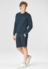Completo in cotone blu navy composto da felpa e pantaloni corti con polsini e vita a coste; presenta un piccolo logo rosso sulla felpa. Sneakers bianche.