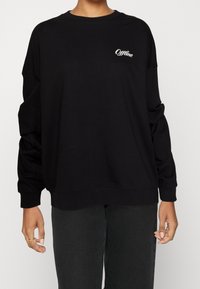 Persona con sudadera negra de manga larga y el texto blanco "Offline" en el pecho, emparejada con pantalones negros, de pie frente a un fondo liso.