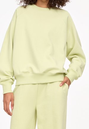 Kvinde iført lysegrøn oversized sweatshirt og matchende bukser, stående med den ene hånd i lommen mod hvid baggrund.