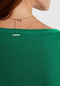 Grüner Strickpullover mit weitem Halsausschnitt und gerippten Kanten. Verfügt über ein kleines Metallschild am Nacken und ein filigranes Kettendetail.