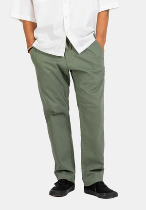 Man wearing olive green casual pants, black sneakers, and a white button-up shirt with hands in pockets.