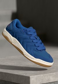 Zilā sneakers ar teksturētu, zamšveidīgu augšu, lentes priekšpusi un baltu vidusdaļu. Iekļauj gumijas zoli un smalku zīmola detaļu.