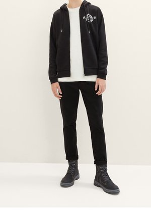 Sudadera negra con cremallera y gráfica blanca, camiseta de color claro debajo, pantalones negros y botas negras con cordones y suelas texturizadas.