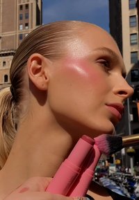 Two pink makeup sticks held near a model's cheek, featuring a shiny blush application and a brush with a pink gradient bristle.