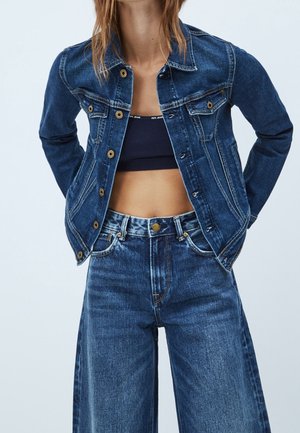 Kvinde iført mørkeblå denimjakke og højtaljede, vidde bukser med sort cropped top, poserer med hænderne bag ryggen.