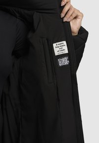 Interior de chaqueta acolchada negra con textura de acolchado, con etiquetas que dicen "Un cálido abrazo hecho de tela" y el logo de la marca en blanco.