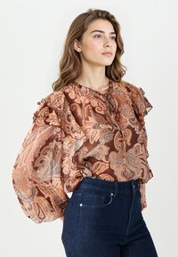 Brązowa bluzka w wzór paisley z falbankami i wiązanym dekoltem, zestawiona z ciemnoniebieskimi dżinsami z wysokim stanem. Przezroczysta faktura materiału.