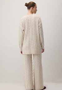 Kaksiosainen neulossetti vaaleassa beessissä, jossa on teksturoituja kaapelikuvioita. Mukana pitkähihainen tunika ja leveälahkeiset housut röyhelöillä.