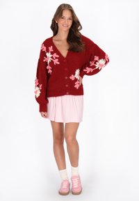 Röd stickad cardigan med broderade rosa blommor, knappstängning, i kombination med en veckad rosa kjol och rosa sneakers. Texturerad och mysig design.