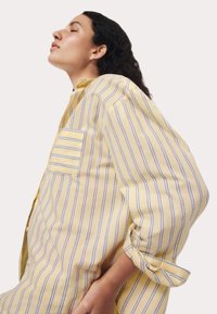 Geel gestreepte shirt met blauwe lijnen, voorzien van een borstzak, lange mouwen en een relaxte pasvorm. De stof lijkt lichtgewicht met een gladde textuur.