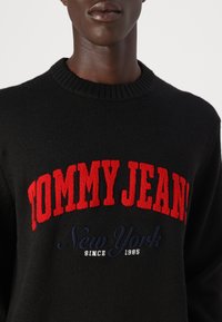 Svart strikket genser med ribbestrikket rund hals. Fronten har stor rød tekst "TOMMY JEAN!" med "New York" og "siden 1985" i marineblått.