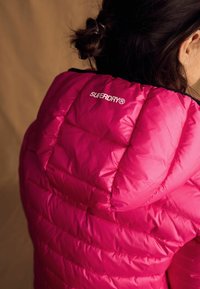Fuchsiová nadýchaná bunda s prešívaným dizajnom a čiernymi akcentmi; obsahuje logo "SUPERDRY" na kapucni. Hladký, lesklý povrch.