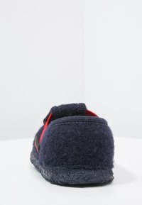 Dunkelblaue Wollhausschuhe mit einer flauschigen Textur, die rote Akzente und eine abgerundete Rückseite aufweisen. Die Sohle ist schwarz und strukturiert für besseren Halt.