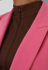 Braunes, geripptes Rollkragenshirt kombiniert mit einem taillierten pinkfarbenen Blazer, der einen sichtbaren Reißverschluss und strukturierte Schultern aufweist und kontrastierende Farben und Texturen betont.